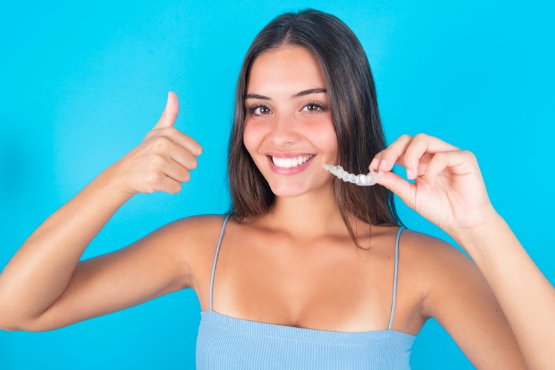A smiling brunette woman holding an Invisalign aligner and giving a thumbs-up